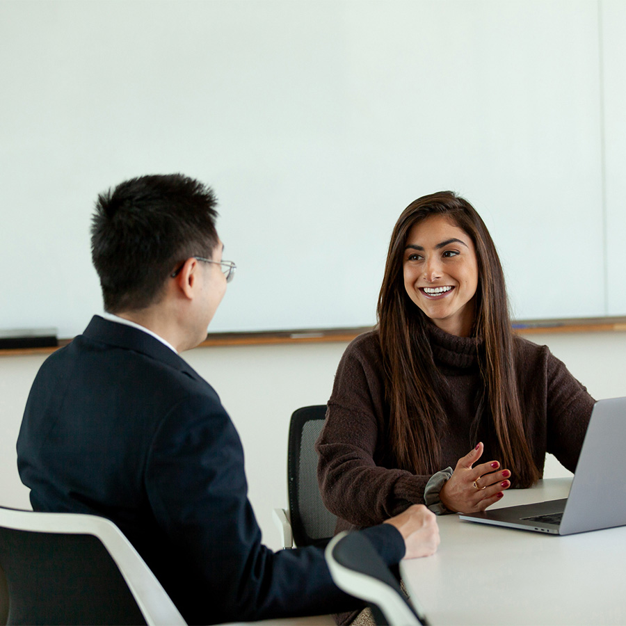 Woman sits at laptop computer while meeting with a man in a suit in a conference room.