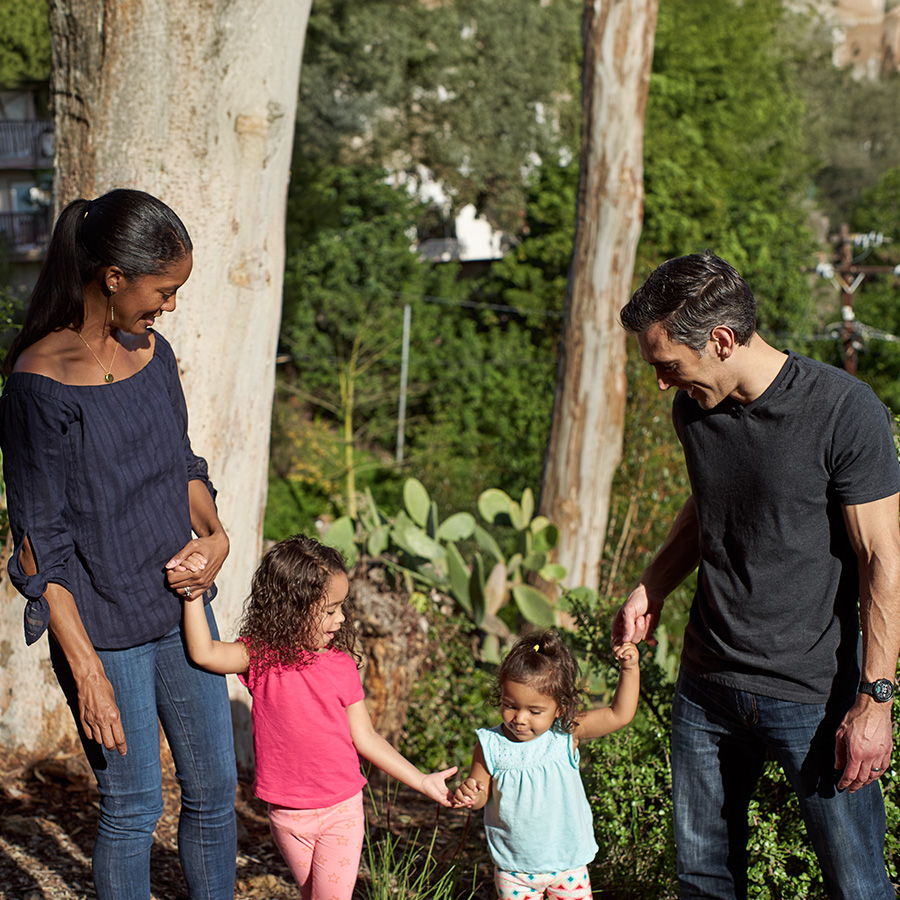 A woman on the left and a man on the right walk along a wooded trail holding hands with two young girls.