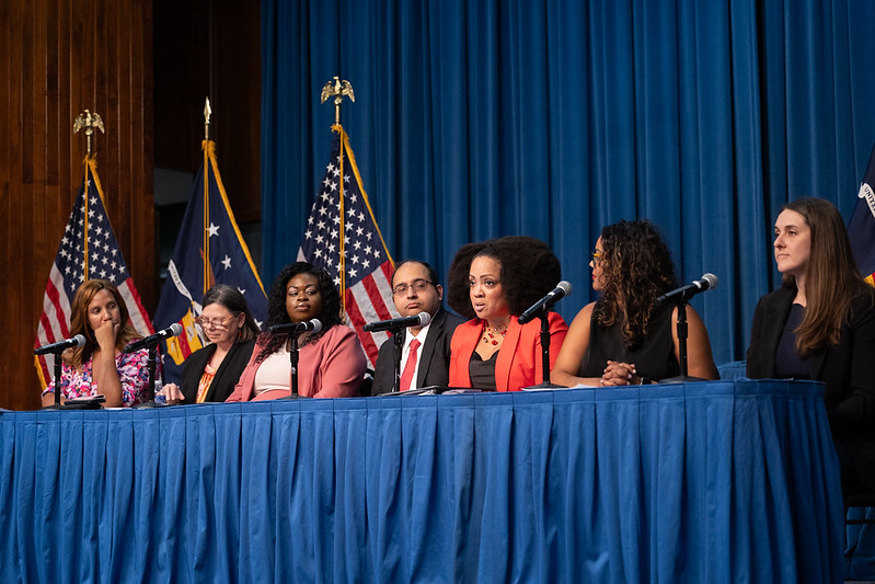 Seven professionally dressed women and men sit on a panel behind a table. American and Labor Department flags are visible on the stage behind them.