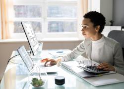 Professionally dressed woman seated at a desk with computers and notebooks.