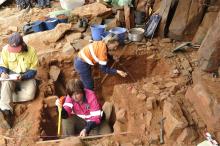 Two women measure the dimensions of a dig-site whilst a man sits beside them taking notes.