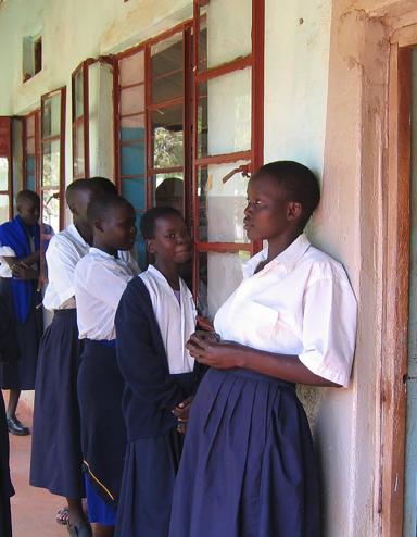 Un groupe de jeunes femmes et d’adolescentes debout sur la véranda d’un bâtiment. La plupart portent des blouses blanches et des jupes bleues, et deux filles portent aussi des chandails bleus. À droite, une porte mène vers une classe remplie d’élèves assises à des pupitres de bois et écrivant dans des carnets. Visibilité masquée.