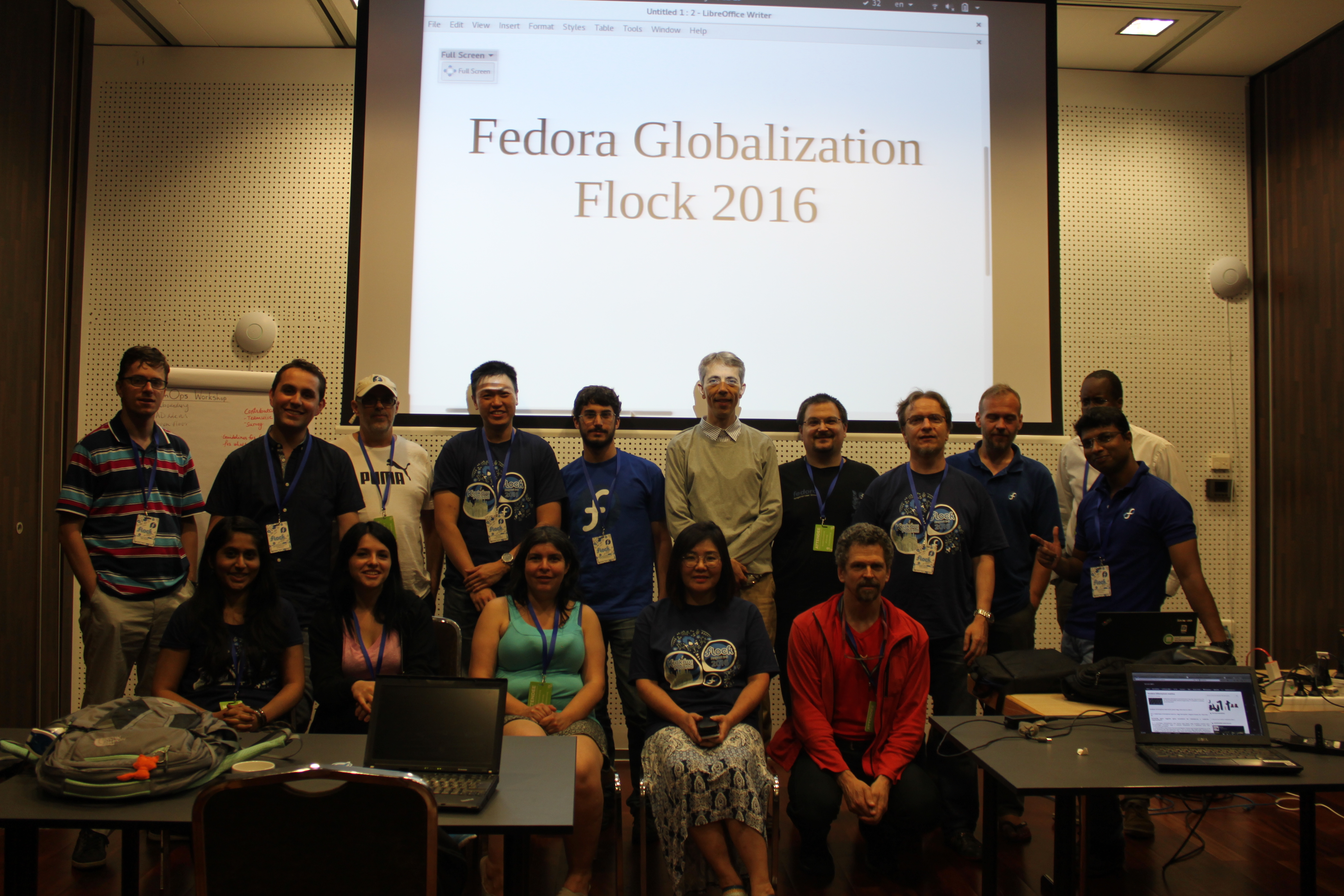 En grupp av cirka 15 personer som alla står för ett foto framför en projektorduk med ett bildspel där det står 'fedora globalization flock 2016