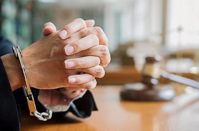 Hands clasped in handcuffs on desk with gavel in background