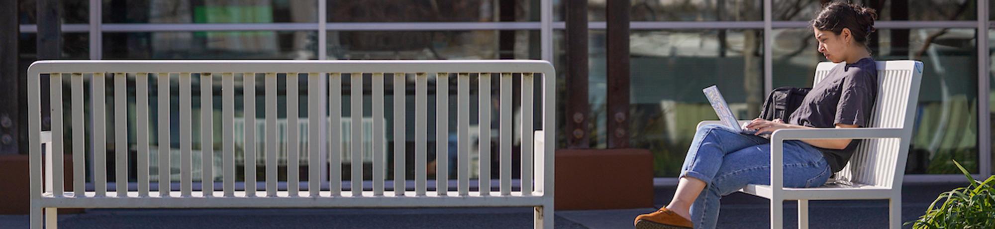 a student wearing jeans and a t-shirt sits on an outdoor bench looking at an open laptop on their knee