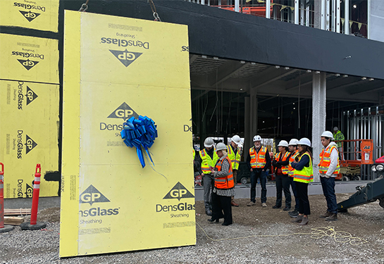 A red crane hoists a yellow wall pane with blue ribbon, watched by people in neon orange vests 