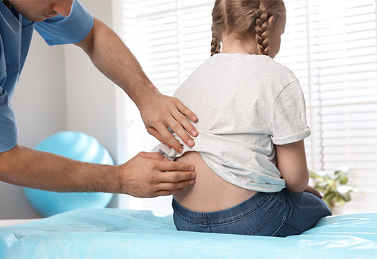 A person wearing blue scrubs and examining the back of a child with blonde braids, wearing white shirt and blue jeans, and sitting on an exam table