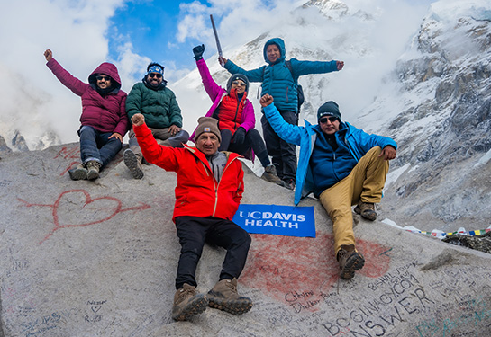 Un hombre con chaqueta roja, guantes negros y gorra color café levanta sus brazos en celebración al frente de una montaña nevada