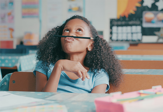 A young girl sits at a school desk, balancing a pencil between her nose and her lip while looking up at the ceiling. 