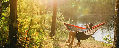 2 people sitting in a hammock in front of a body of water and woods