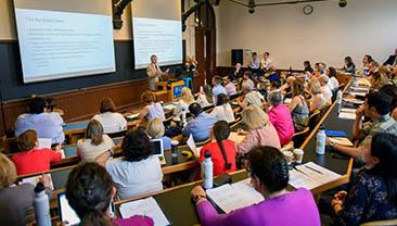 A group of people attending a lecture for the Intensive Bioethics Course, with a man standing at the front of a classroom lecturing, slides projected on dual screens behind him