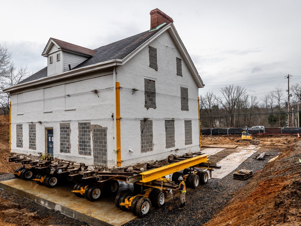 An old white house being moved on a large trailer with wheels.