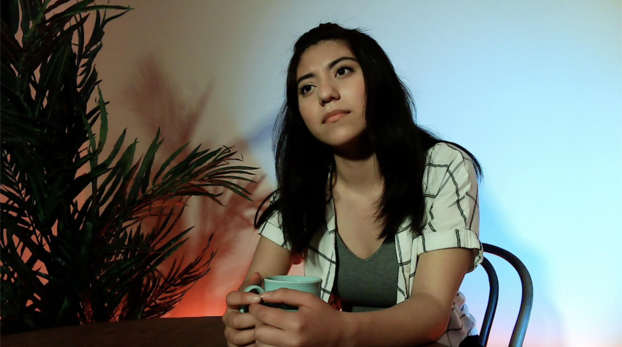 woman sits at a table with a coffee cup in her hand, with blue and red light on the walls behind her