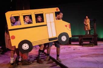 three "Ghost Children" stand on train tracks behind a bus made of paper, looking concerned