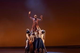 A group of at least seven dancers hold up another dancer, who is holding her arms out at right angles next to her