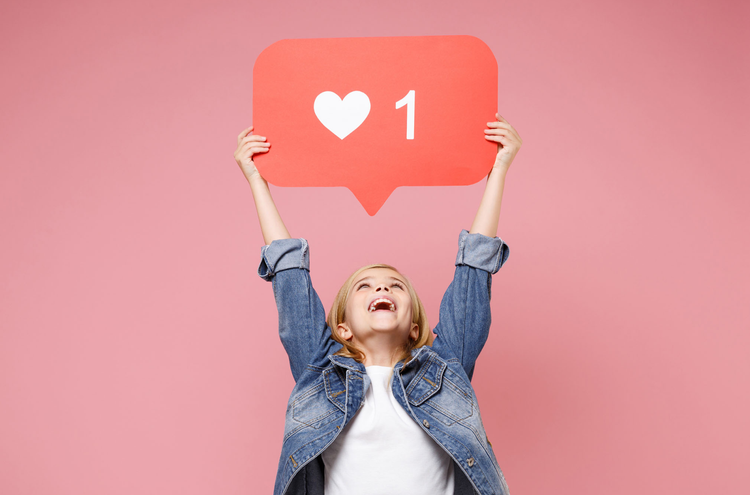 A child holding up a 1 like Instagram notification sign, looking up and smiling, against a pink background