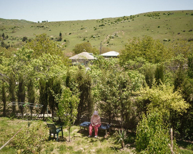 A photo of trees in a field with two little buildings between some of the trees in the back and a person sitting on a bench. in front of the trees with a chair in front of them and a drying rack next to them.