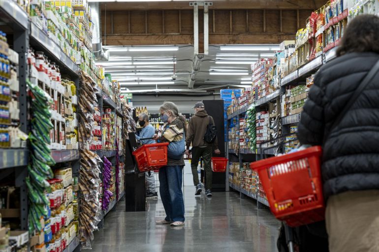 Shoppers in a grocery store aisle, some wearing masks, and carrying red baskets