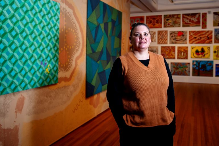 Stacey Edwards. She is standing in front of her art work. She is wearing a black shirt with a brown vest over the top. Her hair is tied back and she has her hands in her pockets