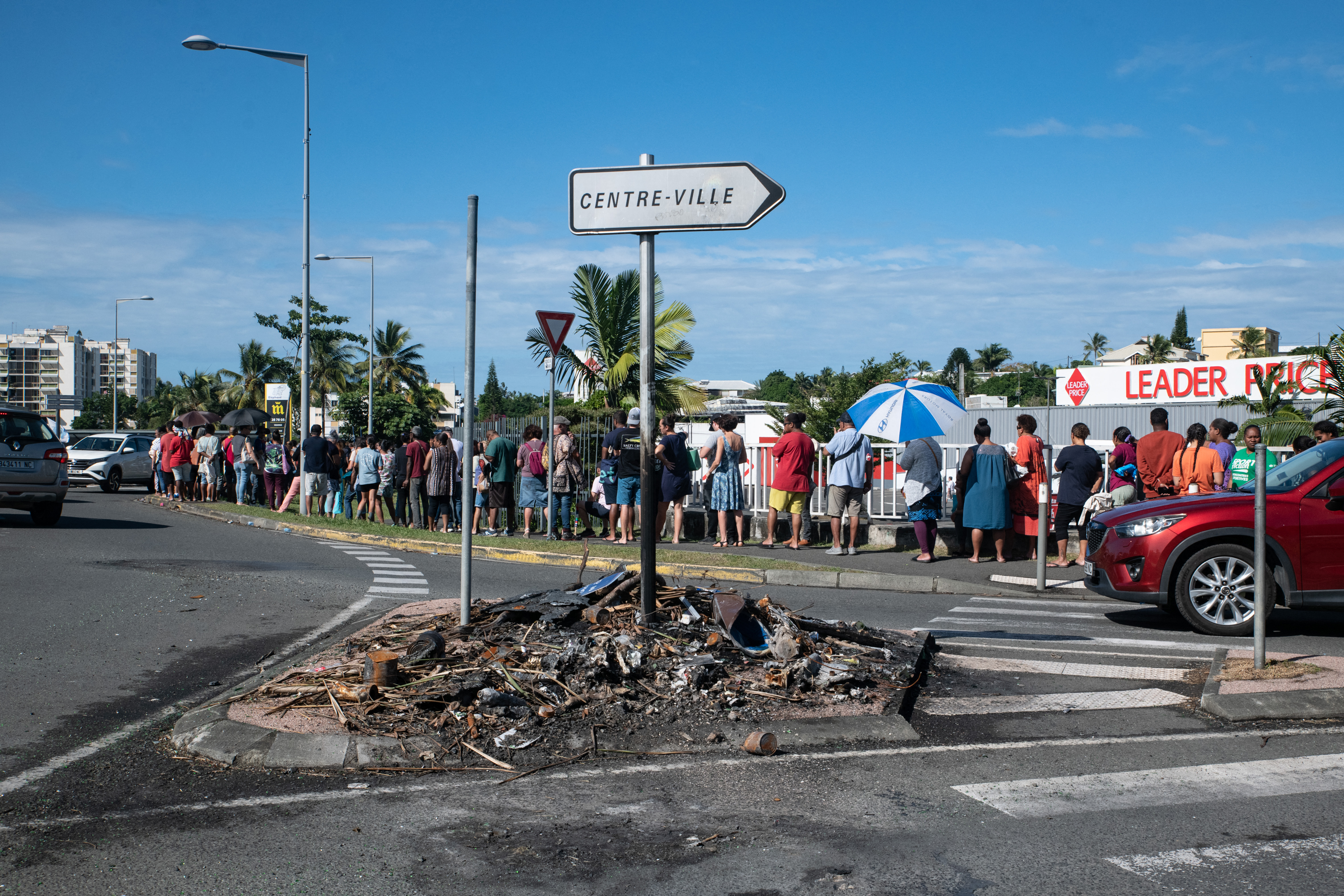 People wait in line to buy provisions from a supermarket as charred items previously set on fire are seen following overnight unrest in the Magenta district of Noumea, France's Pacific territory of New Caledonia, on May 18