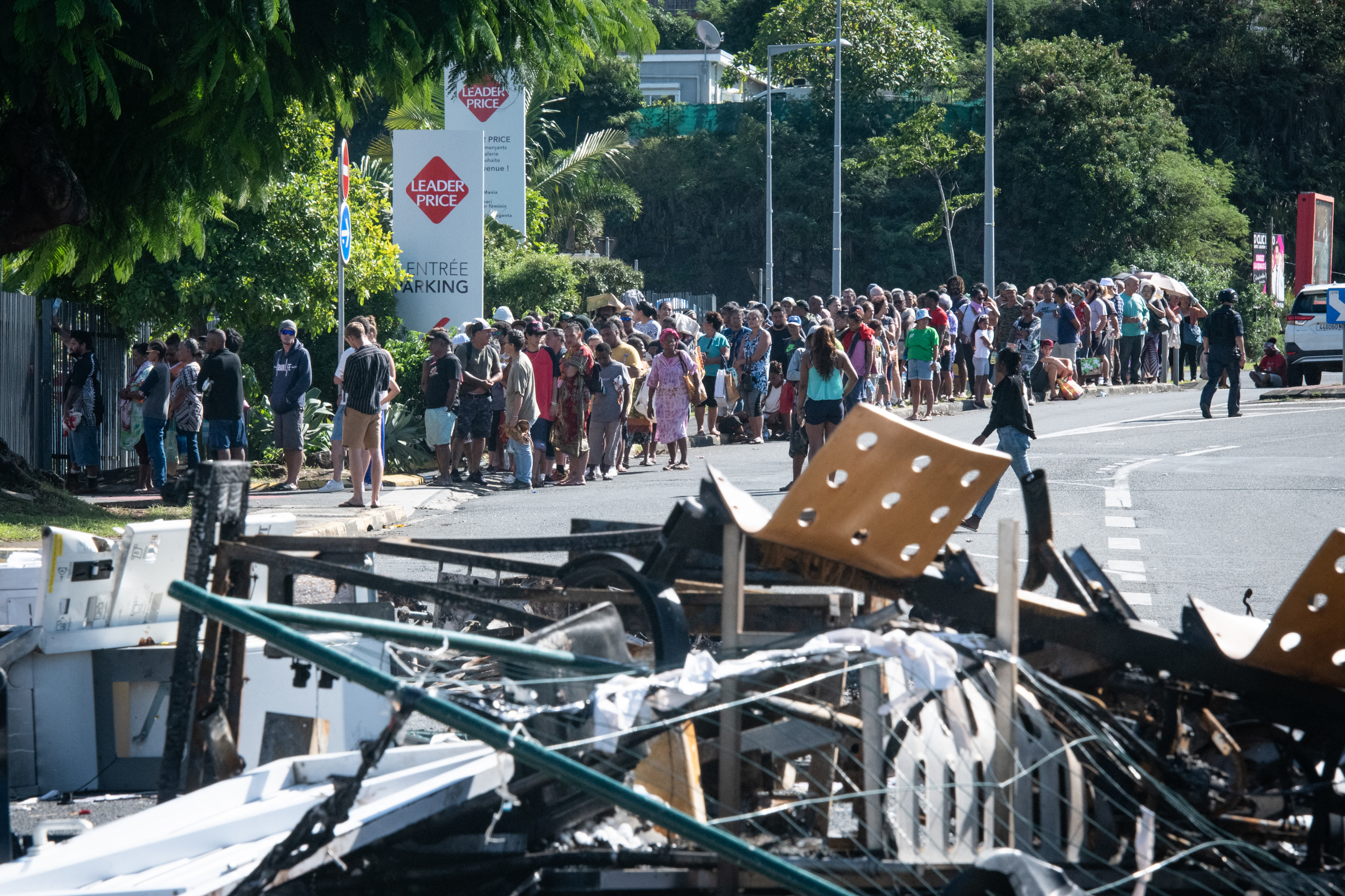 People wait in line to buy provisions from a supermarket along a street blocked by debris and burnt out items following overnight unrest in the Magenta district of Noumea, France's Pacific territory of New Caledonia, on May 18