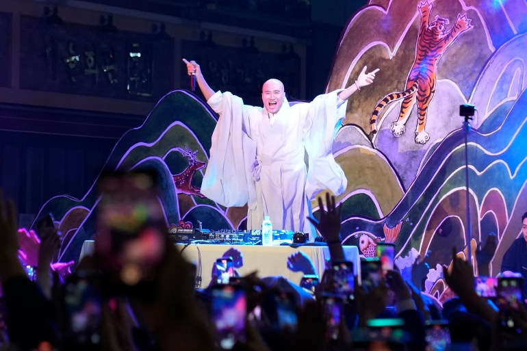 NewJeanNim performing in Seoul. He is wearing a robe and his arms are stretch out. The lighting around him is purple and the crowd are dancing. 