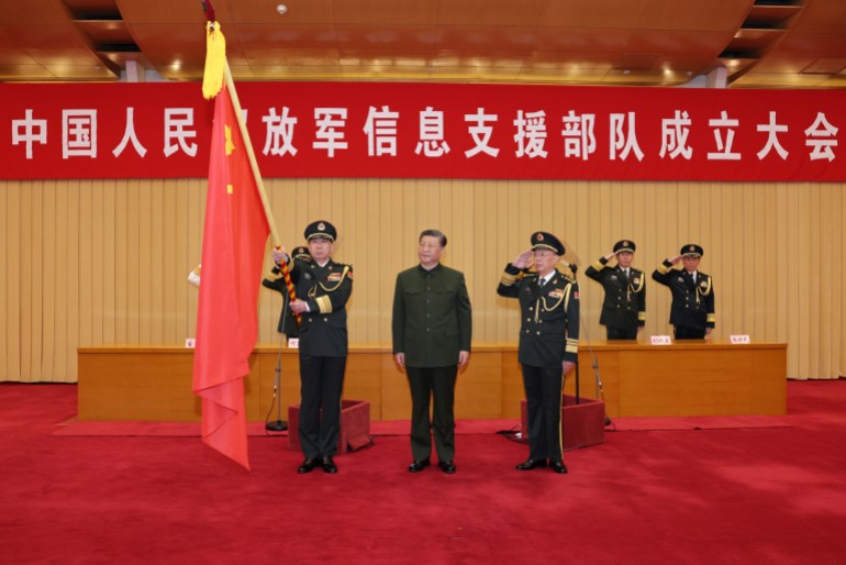Xi Jinping presents a flag to the information support force of the PLA in April. They are on a stage with a red carpet. Xi is between two military officials. There's a banner behind and two soldiers are saluting in the background. 