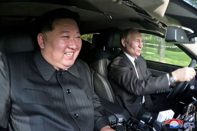 Kim Jong Un grinning as he sits in the passenger seat while Vladimir Putin drives the Aurus