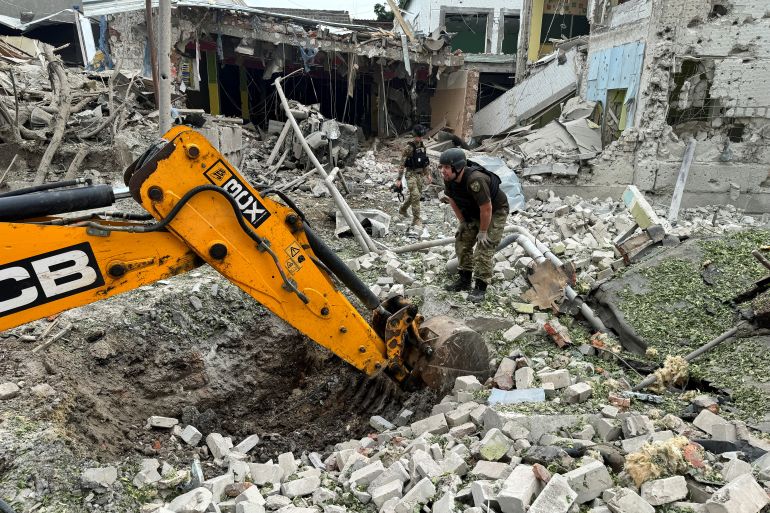 A member of the bomb squad working at the scene of a Russian attack on Kharkiv on Sunday. The officer is standing on a pile of rubble with the destroyed building behind. An excavator is at work.
