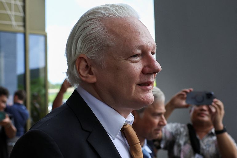 A head and shoulders photo of Julian Assange walking out of court. He is wearing a dark suit, white tie and tobacco-coloured tie. His white hair is combed back. He looks relieved and happy.