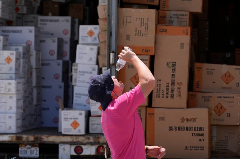 A man in a ballcap and faded red T-shirt pours the last drops of a plastic water bottle into his mouth. Behind him are boxes stacked high.