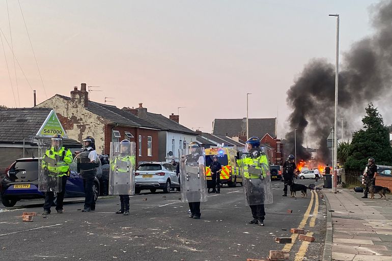 Riot Police standing across a street in Southport. They are are wearing helmets and have transparent shields. Clouds of black smoke are rising into the air behind them. There are bricks lying on the street. Some cars are on fire in the background. There are also officers with dogs.