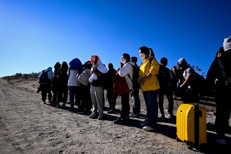 Asylum-seekers from China wait to be processed by U.S. Border Patrol agents after crossing the nearby border with Mexico. They are standing in a line, Some have luggage. One has a bright yellow suitcase