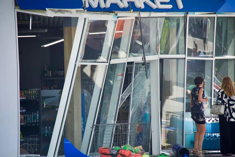 A damaged shop facade after a Russian attack on Dnipro. The window and door frames are bent and sticking outwards. Some of the glass has broken. Two women are standing outside.