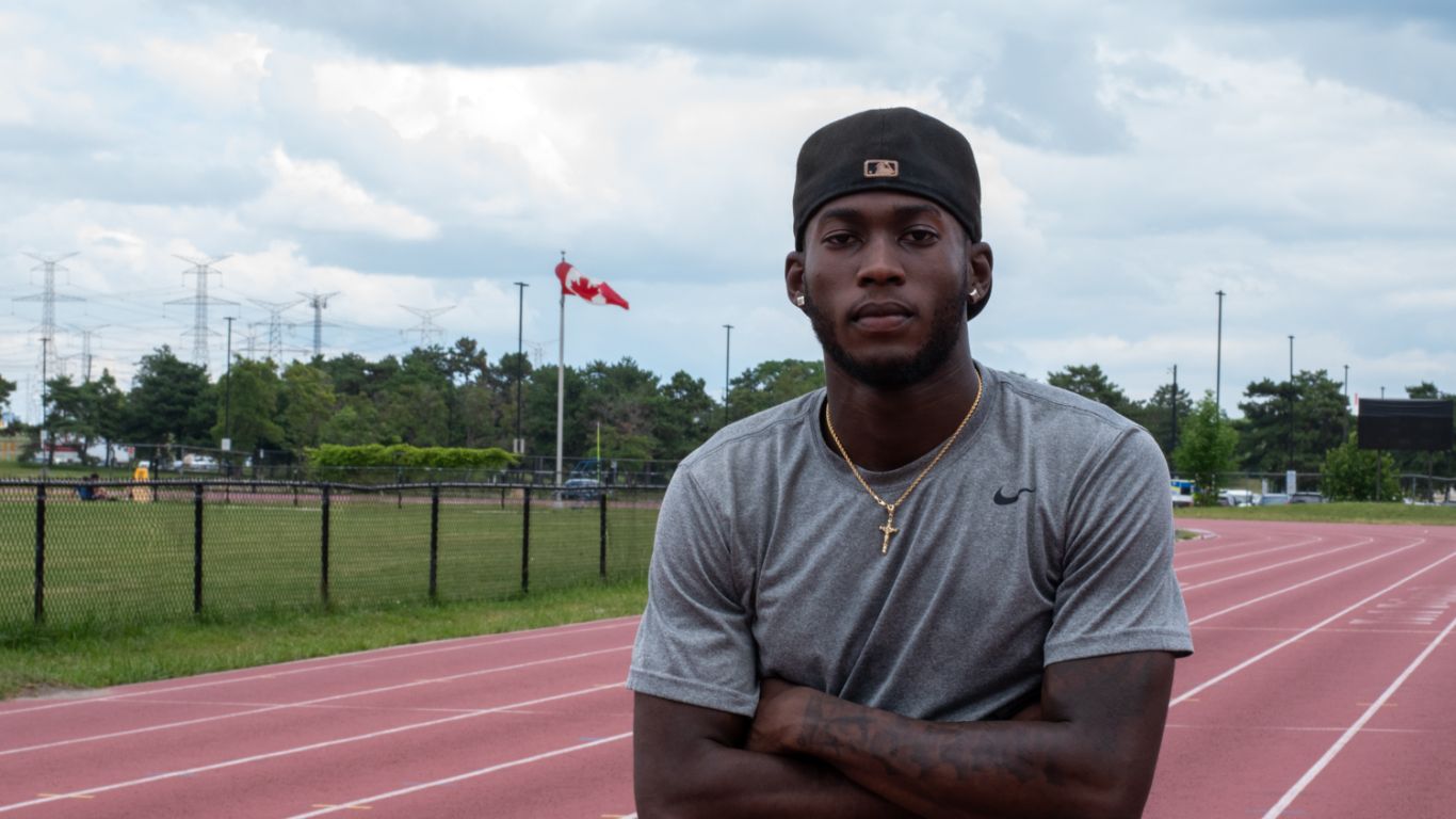 Tamarri stands on an outdoor track cross-armed. Behind him is a flag pole bearing the Canadian flag