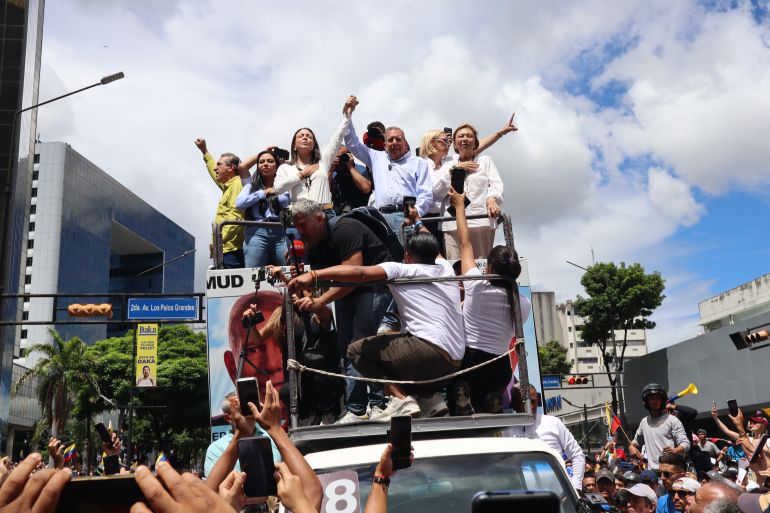 Maria Corina Machado and Edmundo Gonzalez hold hands and lift their arms as they ride atop a truck, surrounded by supporters.