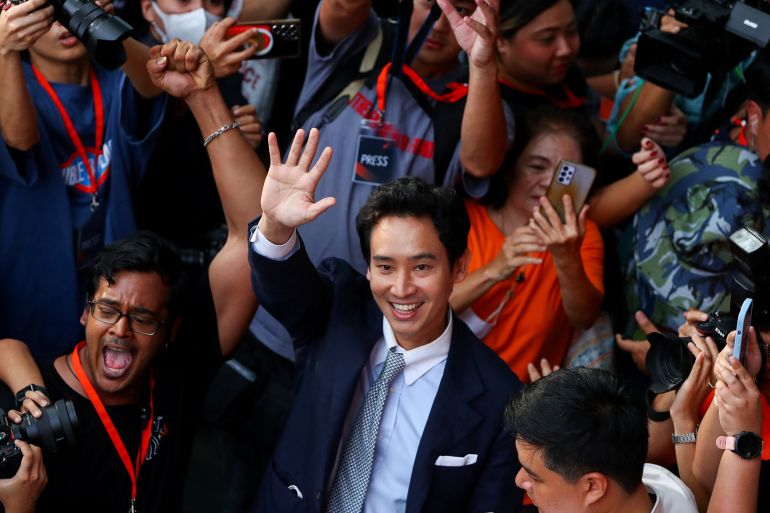 Pita Limjaroenrat waving to supporters. He is wearing a dark suit. pale blue shirt and tie. He is surrounded by photgraphers and supporters. He is smiling.