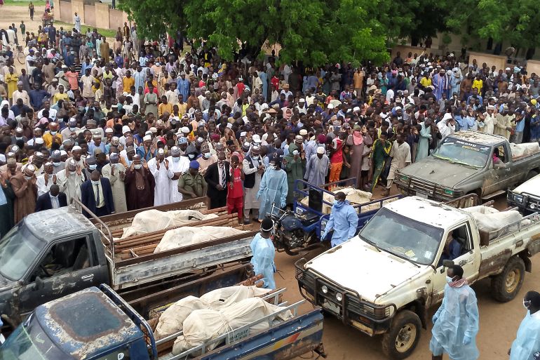 A crowd of people at a funeral for those killed in a suspected Boko Haram attack in northeastern Nigeria. The mourners are crowded towards the back of the picture. There are several pick-up trucks in front with bodies on the back. They are wrapped in white sheets. There are people standing around the trucks. They are wearing protective gowns.