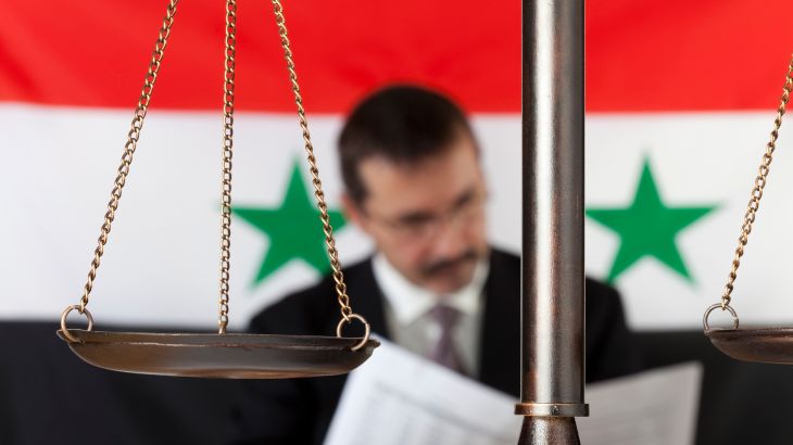 Man sitting by a Syrian flag. Scale of justice in the foreground.