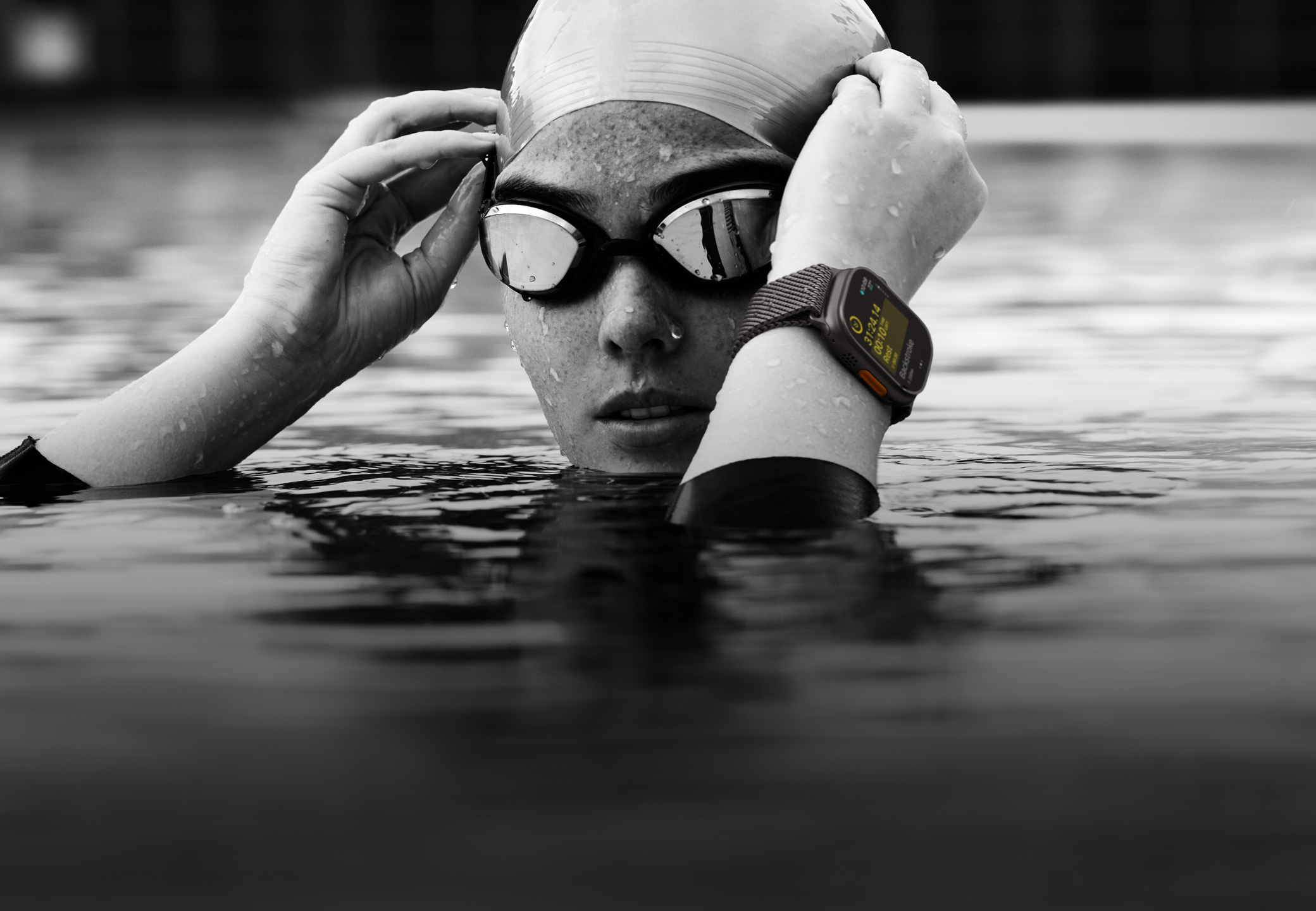 A swimmer’s head and hands above water in a swimming pool wearing Apple Watch Ultra 2 and adjusting goggles.