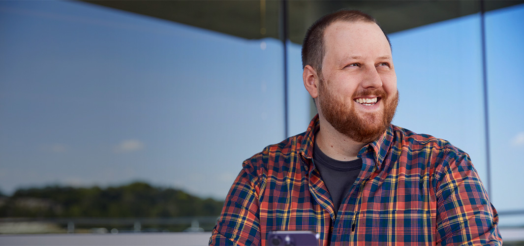 Topher, assis devant un immeuble de bureaux, regarde vers le côté en souriant.