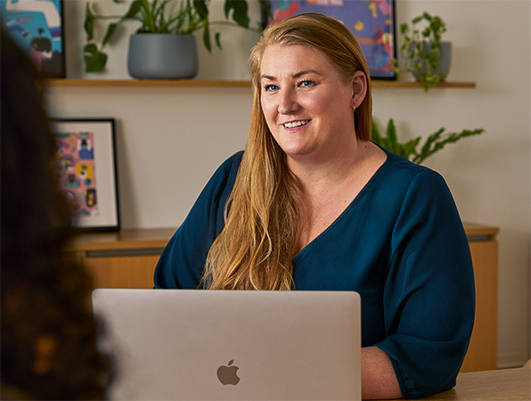 An Apple employee at a table in a common area, using a MacBook while speaking to a colleague.