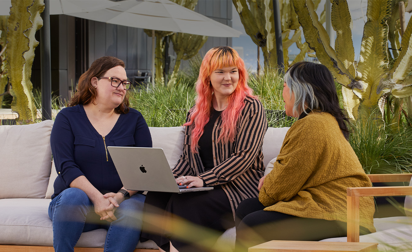 Tres empleadas de Apple, una sostiene una MacBook, están sentadas en un patio exterior.