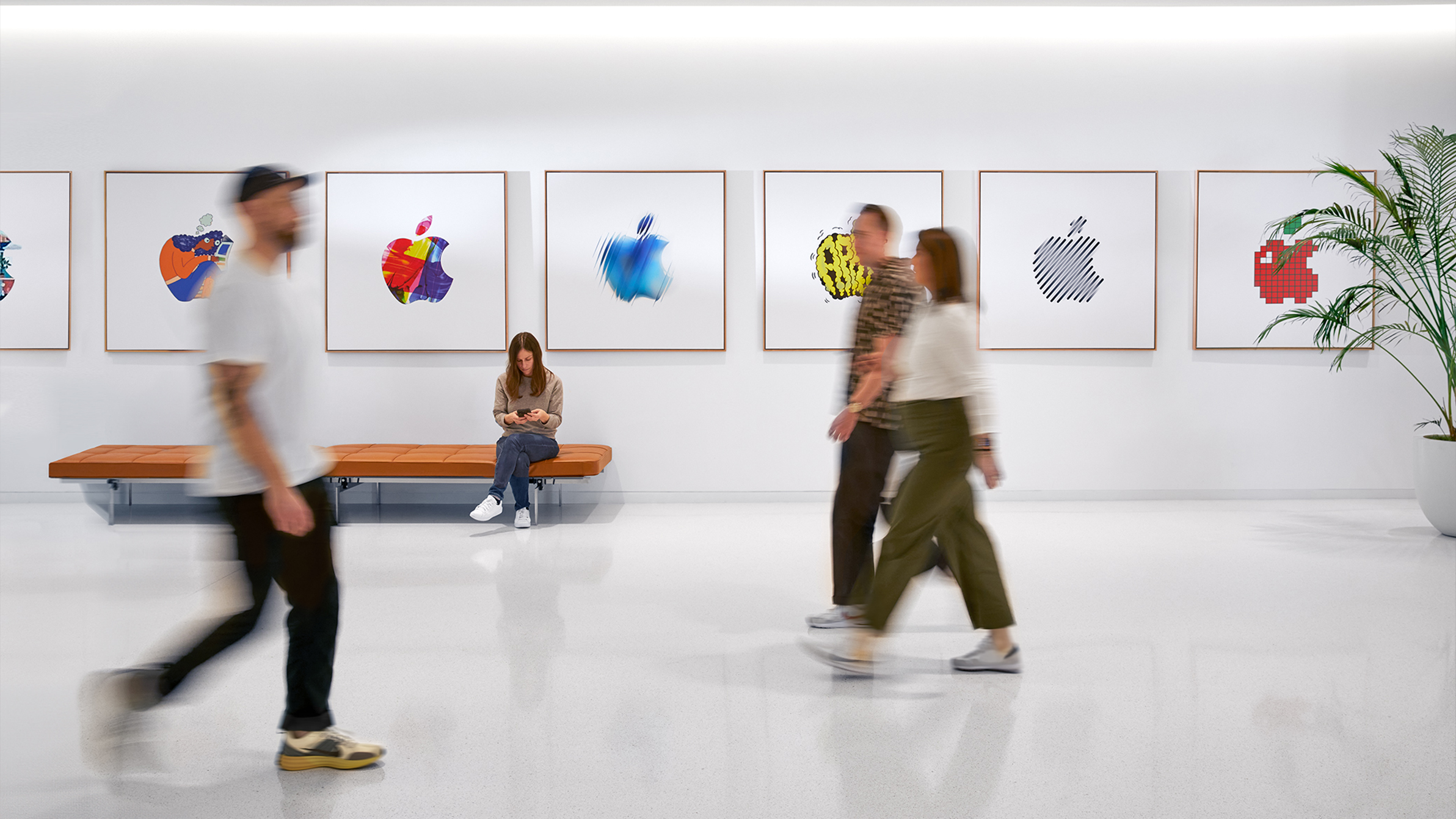 Tres colegas de Apple pasan caminando frente a una pared interior con logotipos coloridos de Apple, una cuarta integrante del equipo está sentada en una banca.