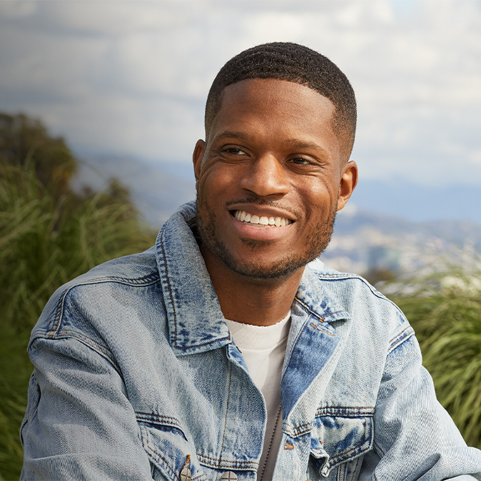 Justin smiling and sitting on an outdoor patio, with sky and trees in the background.