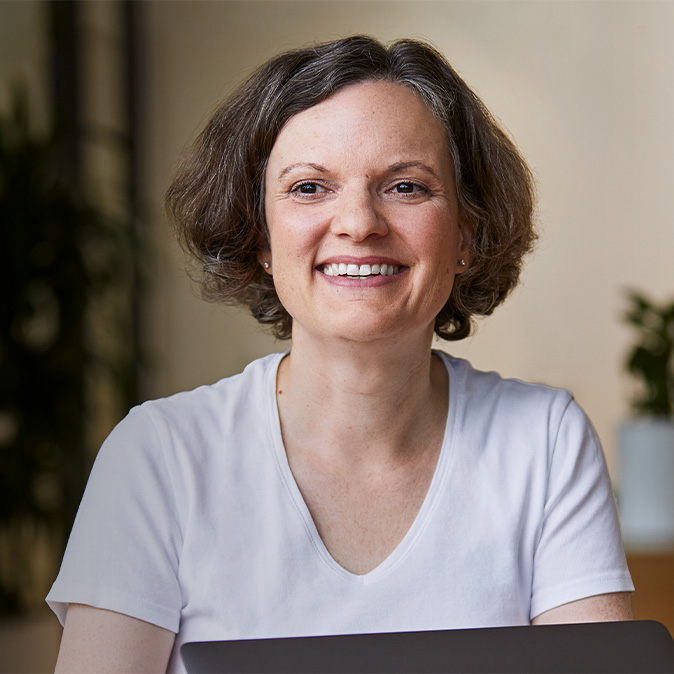 Ruth lächelt und sitzt in einem Raum mit Pflanzen im Hintergrund.