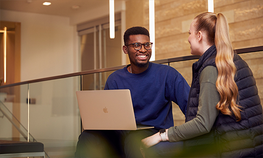 Chidi conversa com uma colega sentado com um MacBook sobre os joelhos.