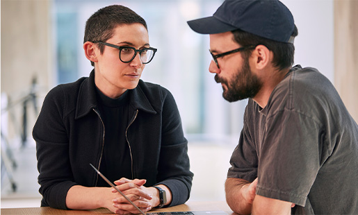 Sophie sidder ved et bord og taler med en kollega, som arbejder på en MacBook.