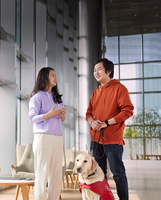 Dos empleados de Apple platican en un área común con mucha luz, uno sostiene la correa de un perro guía.