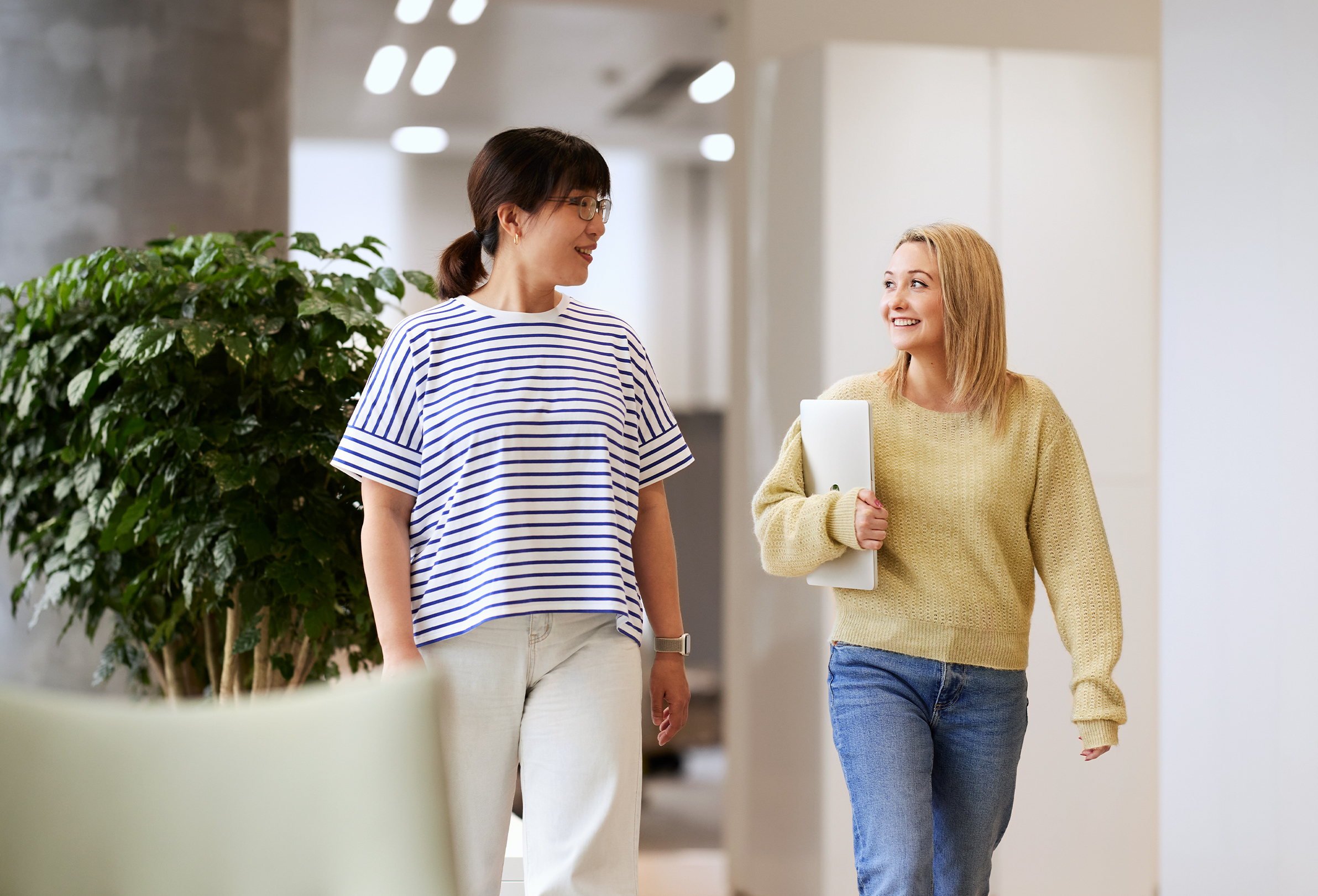 Duas funcionárias conversam e sorriem enquanto caminham por um espaço comum bem iluminado.